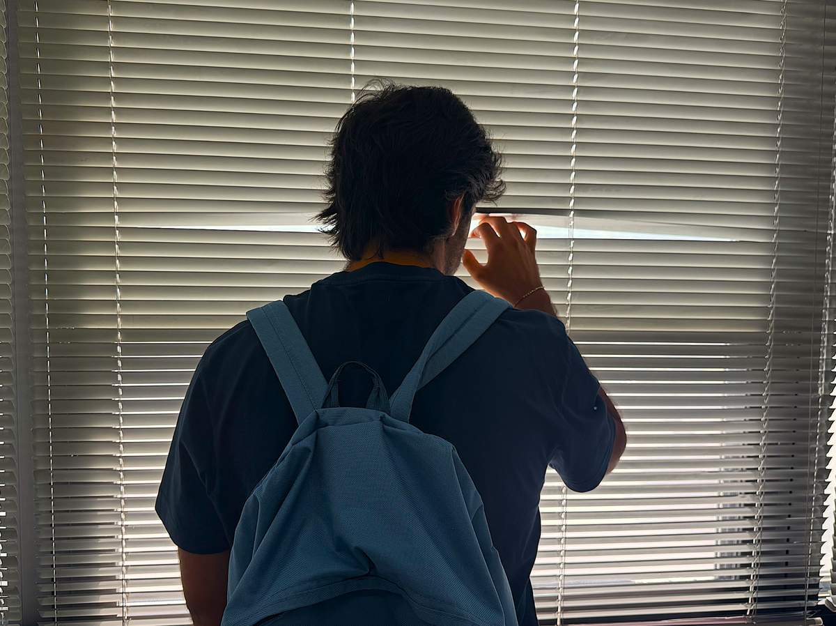 Un ragazzo con lo zaino guarda attraverso le persiane chiuse. Simbolo di solitudine e fragilità adolescenziale.