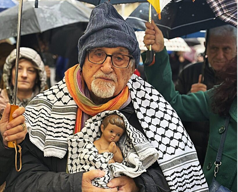 Durante il corteo per Gaza svoltosi a Napoli nella notte di Natale, padre Alex Zanotelli tiene tra le braccia un Gesù Bambino avvolto in una kefiah palestinese, sotto la pioggia, insieme ai manifestanti.