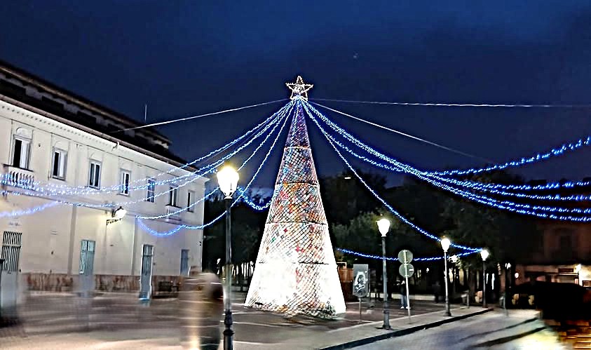 L’albero di Natale all’uncinetto risplende al centro della piazza, avvolto da una rete di luci blu che convergono sulla stella superiore, creando un effetto radiale che abbraccia lo spazio circostante. Se vuoi, posso aggiungere una ver
