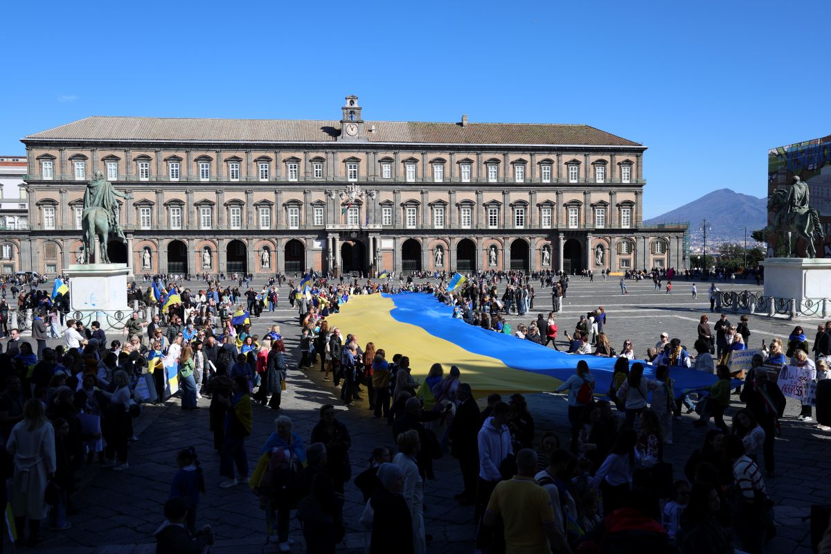 Una veduta ampia del corteo per l’Ucraina a Napoli. In primo piano la grande bandiera blu e gialla portata dai manifestanti; sullo sfondo il Palazzo Reale e il Vesuvio. L’immagine racconta la dimensione collettiva della solidarietà espressa dalla città nel quarto anniversario dell’invasione russa.