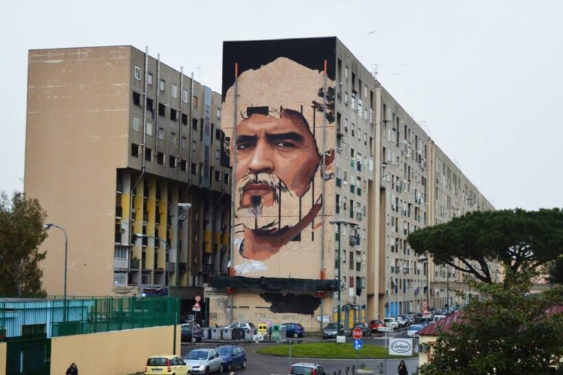 La foto mostra un esteso edificio residenziale in un’area urbana periferica, segnato dalla presenza di un grande murale che domina la facciata.