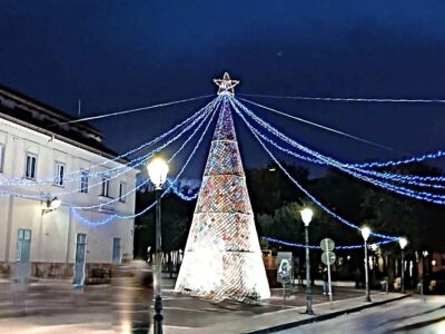 L’albero di Natale all’uncinetto risplende al centro della piazza, avvolto da una rete di luci blu che convergono sulla stella superiore, creando un effetto radiale che abbraccia lo spazio circostante. Se vuoi, posso aggiungere una ver