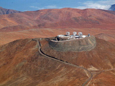 A spectacular aerial view of the ESO Very Large Telescope (VLT) platform, atop Cerro Paranal, in the Chilean Atacama Desert. The VLT consists of an array of four giant 8.2-metre Unit Telescopes (UTs), that can observe together or individually, and four 1.8-metre Auxiliary Telescopes (ATs) dedicated to interferometry. UTs and ATs are visible in the picture. On the right side of the platform is the 2.6-metre VLT Survey Telescope (VST), while on the left edge the Control Building, where astronomers operate the telescopes during the night, is partially visible. The Paranal Observatory is located at 2600 metres altitude, in the Taltal district, some 120 km south of Antofagasta, in the II Region of Chile. In the background at the upper edge of the image is the Pacific Ocean, only 12 km west of the observatory and typically covered by clouds. This is due to the fact that the cold oceanic stream keeps the thermal inversion layer very low, guaranteeing exceptional conditions for ground-based astronomy.