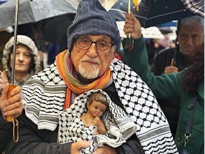 Durante il corteo per Gaza svoltosi a Napoli nella notte di Natale, padre Alex Zanotelli tiene tra le braccia un Gesù Bambino avvolto in una kefiah palestinese, sotto la pioggia, insieme ai manifestanti.