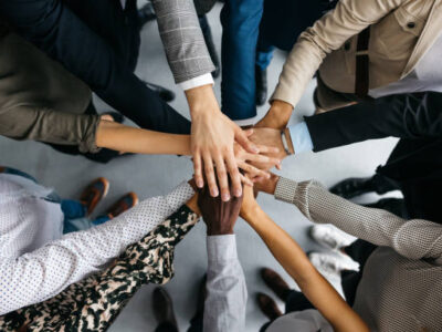 A high-angle shot of a group of male and female colleagues putting their hands together in an office. They are dressed in fashionable business clothes. Their faces are not visible, only their arms. Horizontal daylight indoor photo.
