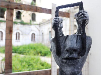 Una scultura contemporanea raffigurante un volto umano che solleva una forma simile a una porta o a una cornice. L’opera, collocata all’interno dell’ex complesso manicomiale di Aversa, suggerisce simbolicamente un gesto di apertura e di cambiamento di prospettiva, invitando a riflettere sul tema dello sguardo, della memoria e della trasformazione.
