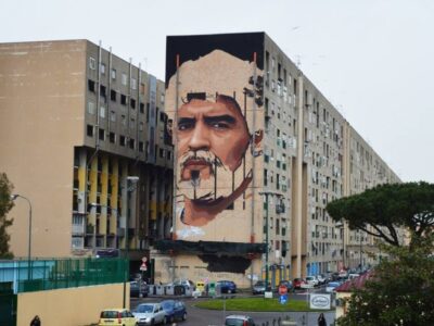 La foto mostra un esteso edificio residenziale in un’area urbana periferica, segnato dalla presenza di un grande murale che domina la facciata.